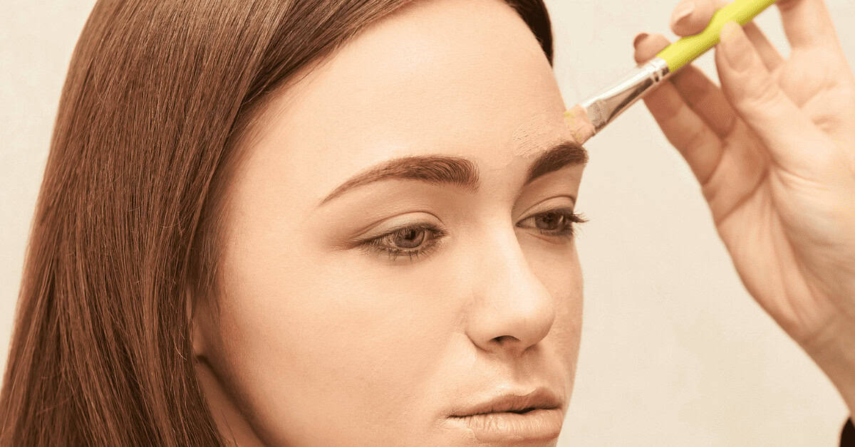 USE CONCEALER TO CLEAN UP MISTAKES Soft focused close-up of a woman receiving eye makeup application, with a brush applying concealer or eyeshadow to her brow area, highlighting beauty, makeup, and eye enhancement techniques.