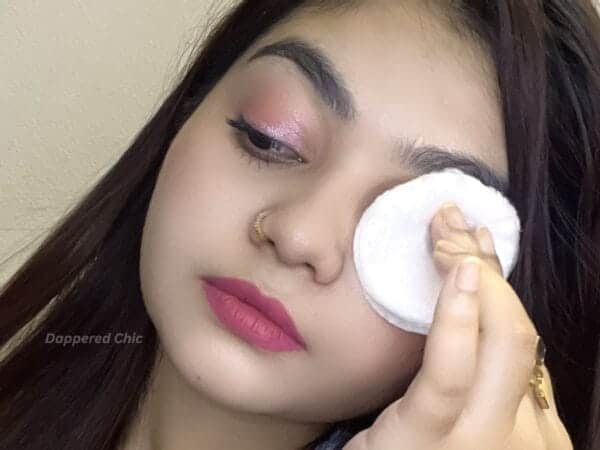 Soft pink eye makeup with subtle shimmer enhances her eyes, complemented by bold, matte pink lipstick. The model uses a cotton pad to remove makeup, showcasing skincare and beauty routine, emphasizing flawless skin and well-groomed eyebrows.